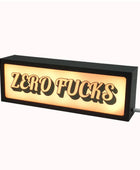 A rectangular black LED sign with the text 'ZERO FUCKS' in raised black letters against a glowing warm white background, shown on a plain light background with a power cord extending from the side.
