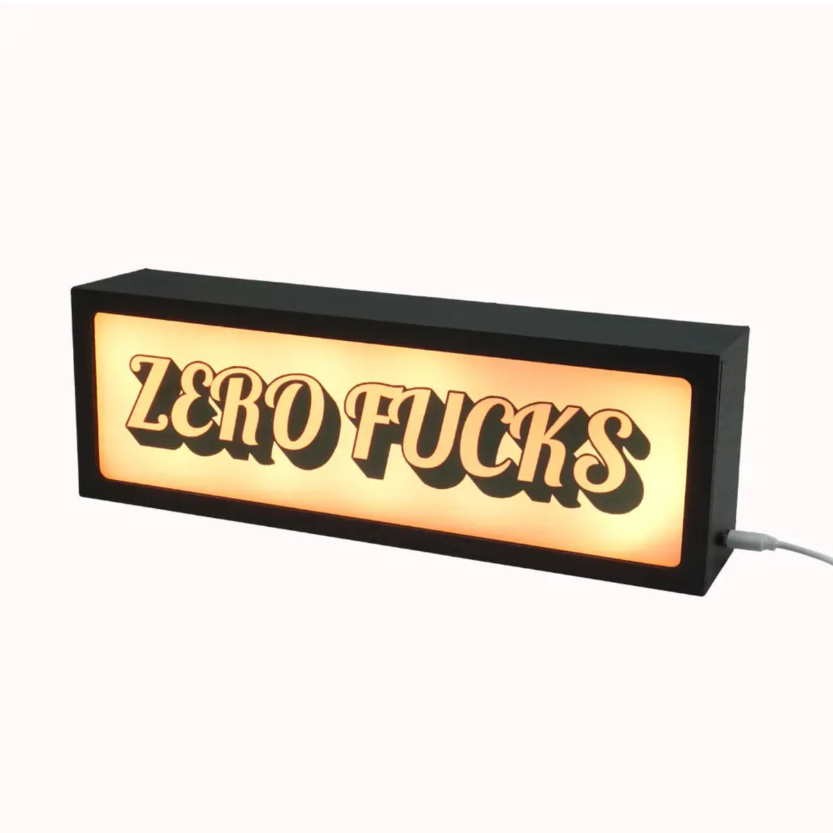 A rectangular black LED sign with the text 'ZERO FUCKS' in raised black letters against a glowing warm white background, shown on a plain light background with a power cord extending from the side.