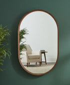 Oval mirror with wooden frame on a green wall reflecting a room with a chair and table.