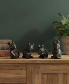 Four small black animal figurines on a wooden surface with a plant and books in the background.