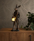 Statue of a rabbit holding a lantern on a wooden surface with a plant in the background