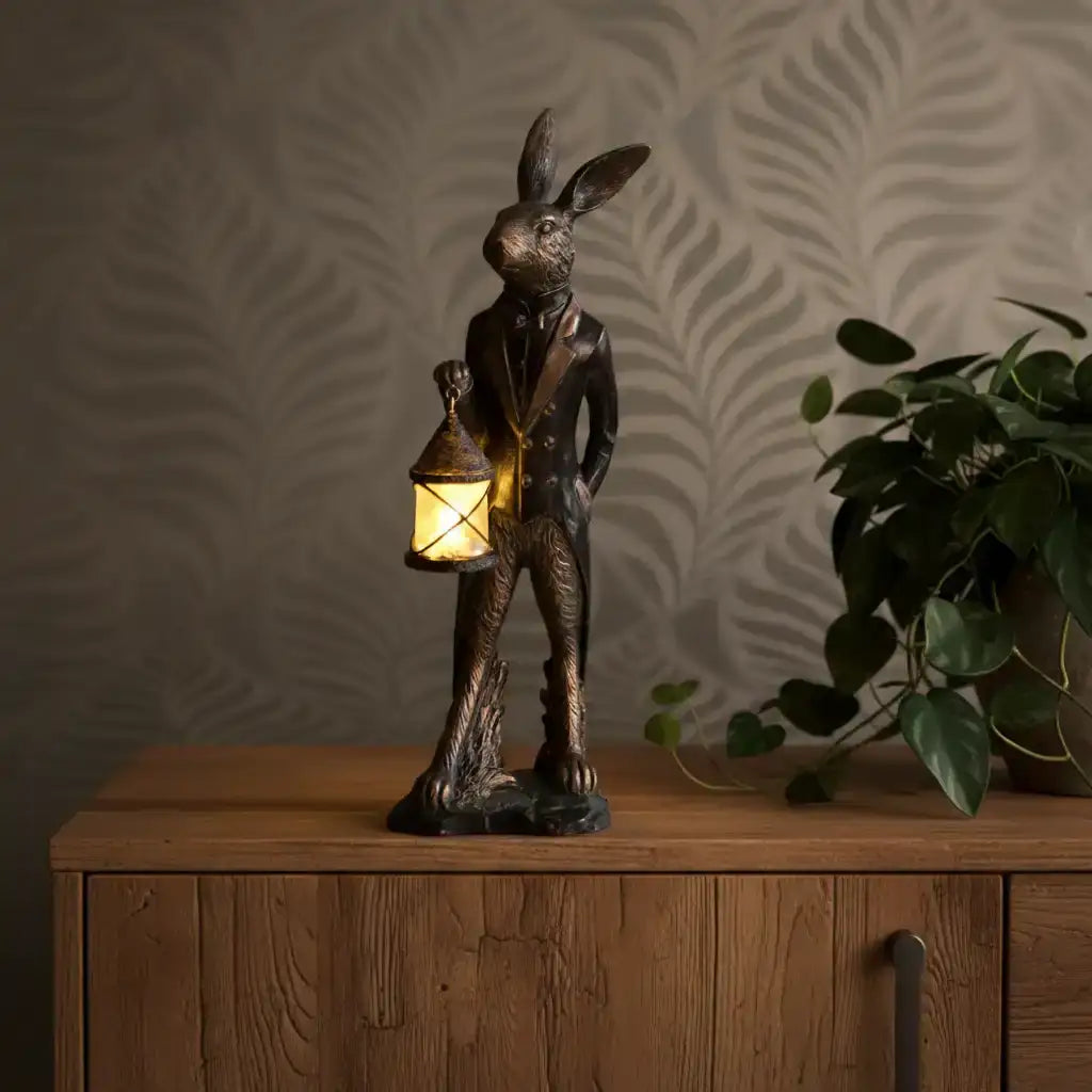 Statue of a rabbit holding a lantern on a wooden surface with a plant in the background