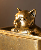 A close-up of a small metallic cat figurine with closed eyes, resting on top of a gold-toned rectangular surface against a dark background.