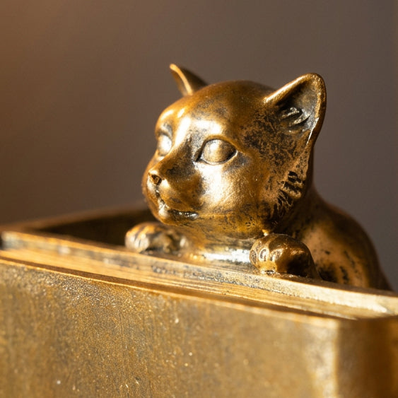 A close-up of a small metallic cat figurine with closed eyes, resting on top of a gold-toned rectangular surface against a dark background.