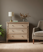 Wooden dresser with a lamp and vase next to a gray armchair in a room with a plant.