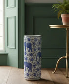 Tall cylindrical container with blue elephant design on a wooden floor next to a green wall and small table with a plant.