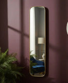 Tall vertical mirror with gold frame on a purple wall, reflecting a room with a lamp and plant.