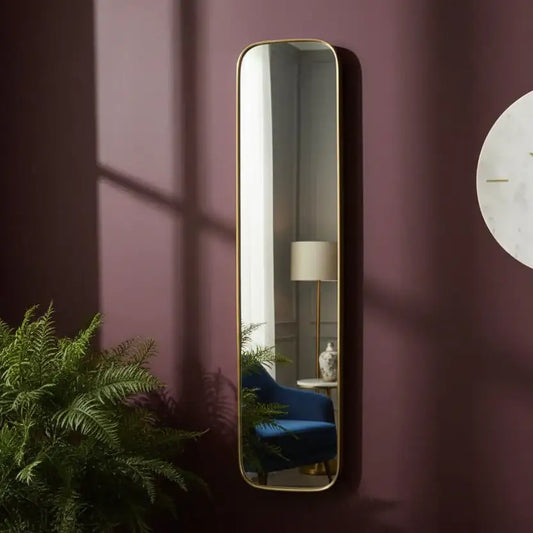Tall vertical mirror with gold frame on a purple wall, reflecting a room with a lamp and plant.