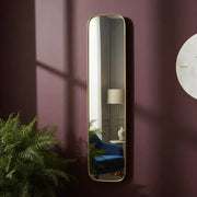 Tall vertical mirror with gold frame on a purple wall, reflecting a room with a lamp and plant.