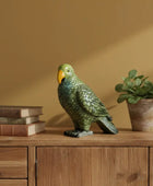 Green parrot figurine on a wooden surface with books and a plant against a beige wall.