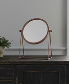 Round stand-up mirror with bronze frame on a dark wooden surface, with a plant beside it.