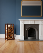 White fireplace against a blue wall with a mirror above it and firewood storage on the left.