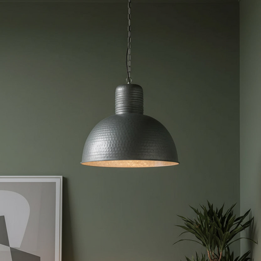 A dark gray metal pendant light with a textured dome shade hanging against a muted green wall, with a framed print and a plant partially visible in the room.