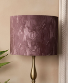 A table lamp with a cylindrical drum shade in a textured, marbled plum-purple fabric, mounted on a polished brass-finished metal base, set against a beige wall.