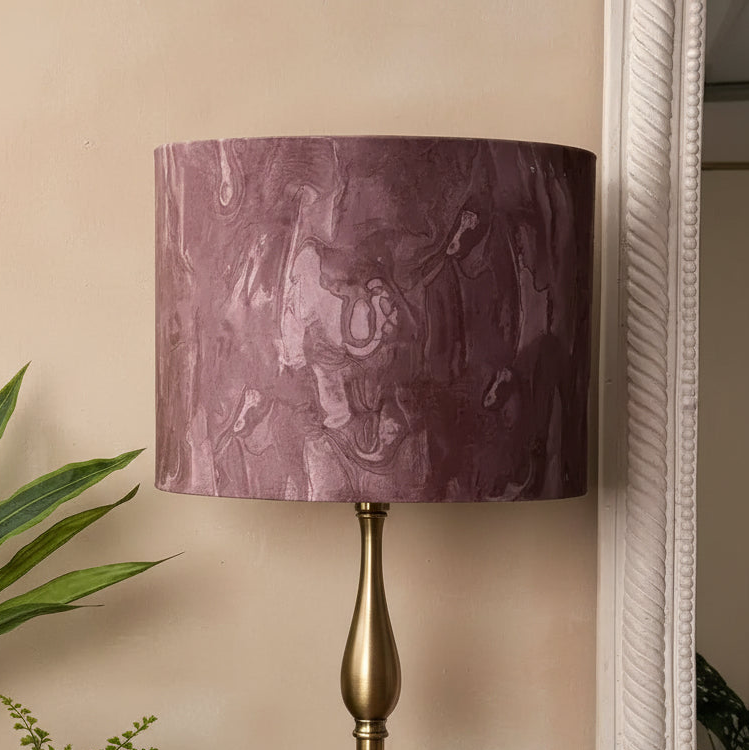 A table lamp with a cylindrical drum shade in a textured, marbled plum-purple fabric, mounted on a polished brass-finished metal base, set against a beige wall.