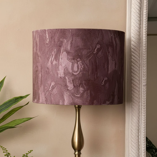 A table lamp with a cylindrical drum shade in a textured, marbled plum-purple fabric, mounted on a polished brass-finished metal base, set against a beige wall.