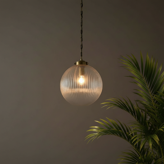 Glass pendant light with gold fixture hanging against a dark background with a plant to the right.