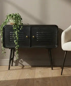 Black side table with gold handles next to a beige armchair and plant on a wooden floor.
