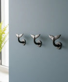 Three whale tail-shaped wall hooks on a gray wall with a plant in the corner.