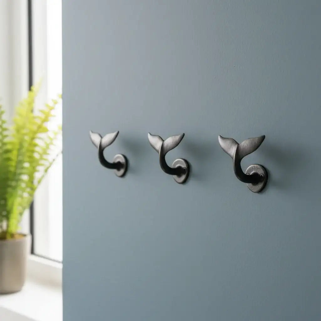 Three whale tail-shaped wall hooks on a gray wall with a plant in the corner.