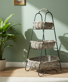 Three-tiered metal stand with wicker baskets against a green wall with a plant on the left.