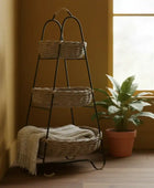 Three-tiered woven basket stand with a blanket against a yellow wall with a window.