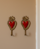 A pair of vintage-style wall hooks shaped like sacred hearts, featuring red enamel hearts set in ornate gold-tone metal frames, mounted on a beige wall.