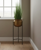 A tall green ornamental grass in a round metallic bronze planter on a black metal stand, placed in a bright room corner with a window, beige walls, and light wood flooring.