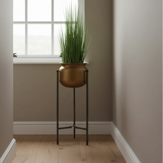 A tall green ornamental grass in a round metallic bronze planter on a black metal stand, placed in a bright room corner with a window, beige walls, and light wood flooring.