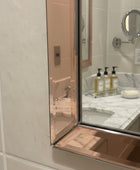 A close-up of a mirror with a metallic rose gold frame installed on a tiled wall, with a hand holding a smartphone visible in the reflection.