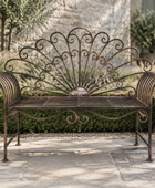 An ornate wrought iron garden bench with a fan-shaped backrest and curved armrests, placed on a stone patio in a landscaped garden with hedges and plants.