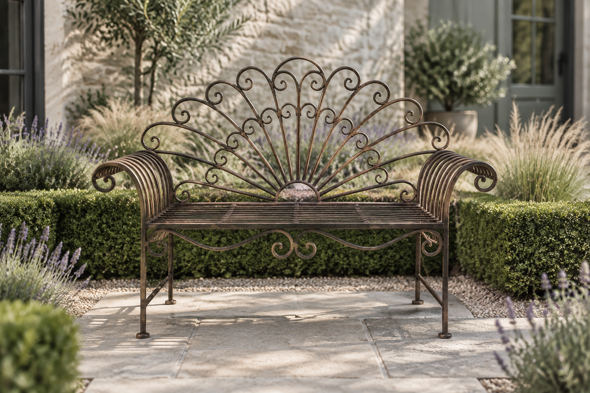An ornate wrought iron garden bench with a fan-shaped backrest and curved armrests, placed on a stone patio in a landscaped garden with hedges and plants.