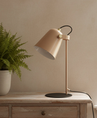 A modern table lamp with a warm beige shade and stem, a black circular base, and a black cord, styled on a rustic wooden chest next to a potted fern against a beige wall.