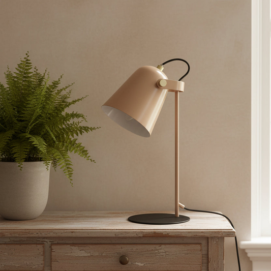 A modern table lamp with a warm beige shade and stem, a black circular base, and a black cord, styled on a rustic wooden chest next to a potted fern against a beige wall.