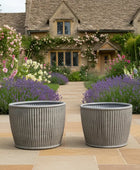 Two gray planters in front of a charming house with blooming flowers.