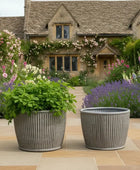Two gray planters with greenery in front of a charming house with floral decorations.