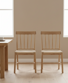 Two wooden chairs in a room with a table and windows.