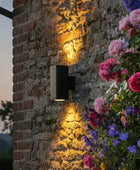 Wall-mounted outdoor light fixture on a stone wall with flowers in the foreground
