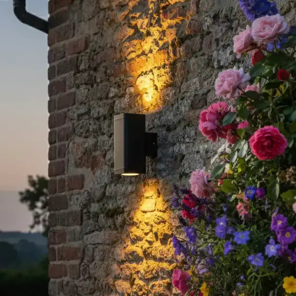 Wall-mounted outdoor light fixture on a stone wall with flowers in the foreground