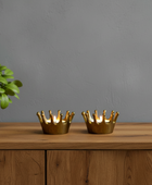 Two gold crown-shaped candle holders on a wooden surface with a plant in the background.