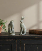 Two stone rabbit sculptures on a wooden surface with a plant and book in the background.