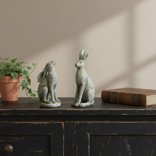 Two stone rabbit sculptures on a wooden surface with a plant and book in the background.