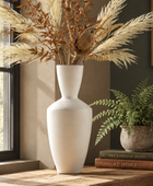 A tall matte white ceramic vase filled with dried pampas grass and brown branches, displayed on a wooden table in a styled interior next to a potted fern and books.