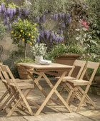 Wooden outdoor dining set with chairs on a wooden deck surrounded by flowers and plants.